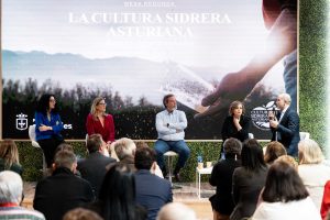 Personas ubicadas a modo de mesa redonda, con gran pantalla detrás y público asistente