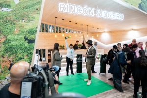 Persona escanciando en zona sidrera del stand de Asturias con público y prensa